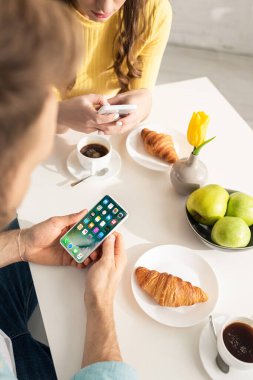KYIV, UKRAINE - 21 Şubat 2020: Akıllı telefonu kız arkadaşı ve mutfağında kahvaltısı olan iPhone ekranlı bir adamın seçici odak noktası 