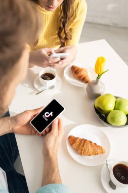 KYIV, UKRAINE - 21 Şubat 2020: Kadınların yanında TikTok uygulaması ve mutfakta kahvaltısı olan akıllı telefonu tutan erkeğin seçici odak noktası 