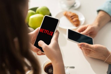 KYIV, UKRAINE - 21 Şubat 2020: Netflix uygulamalı akıllı telefon kullanan kız odaklı erkek arkadaş ve kahvaltı masasında 