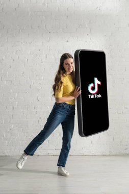 KYIV, UKRAINE - 21 Şubat 2020: TikTok uygulamalı akıllı telefonun büyük modelini tutarken gülümseyen ve kameraya bakan güzel kız