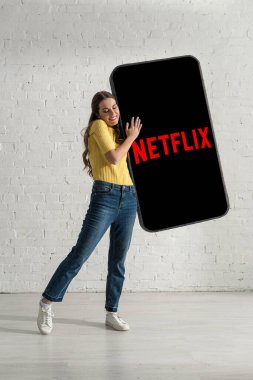 KYIV, UKRAINE - 21 Şubat 2020: Gülümseyen kız evde netflix uygulaması ile akıllı telefonun büyük modelini kucaklıyor 