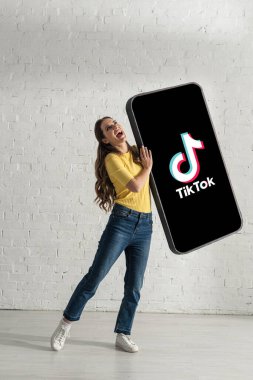 KYIV, UKRAINE - 21 Şubat 2020: Beyaz tuğla duvarın yanında TikTok uygulaması olan akıllı telefonun büyük bir modelini tutarken gülen çekici kadın