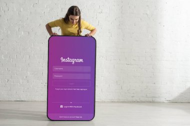 KYIV, UKRAINE - 21 Şubat 2020: Beyaz tuğla duvarın yanında instagram uygulaması olan akıllı telefonun büyük modelinin yanında duran genç kadın