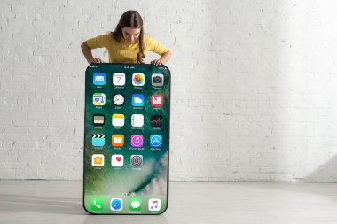 KYIV, UKRAINE - 21 Şubat 2020: Beyaz tuğla duvarın yanında iPhone ekranı olan büyük akıllı telefon modeline bakan kız