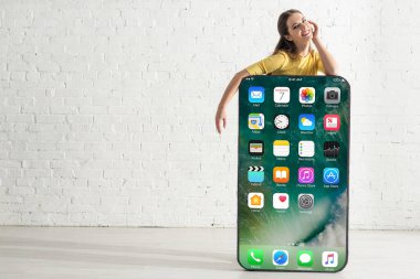 KYIV, UKRAINE - 21 Şubat 2020: İphone ekranlı akıllı telefonun büyük modelinin yanında dururken eli yanağına yakın gülümseyen güzel kadın