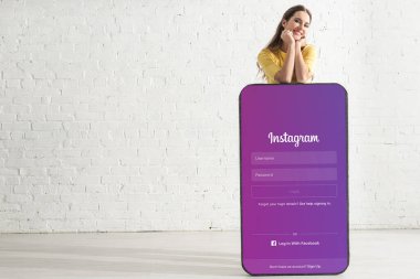 KYIV, UKRAINE - 21 Şubat 2020: Evinde instagram uygulaması olan akıllı telefonun büyük modelinin yanında kameraya gülümseyen genç kadın 