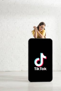 KYIV, UKRAINE - 21 Şubat 2020: Gülen kadın beyaz tuğla duvarın yanında TikTok uygulaması olan akıllı telefonun büyük modeline bakıyor