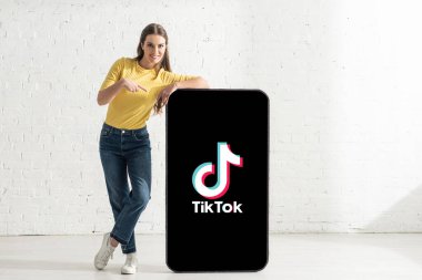 KYIV, UKRAINE - 21 Şubat 2020: TikTok uygulamalı akıllı telefonun büyük modelini işaret ederken kameraya bakan mutlu kadın