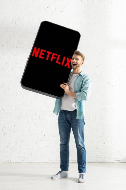 KYIV, UKRAINE - 21 Şubat 2020: Beyaz tuğla duvarın yanında netflix uygulamalı akıllı telefonun modelini tutan heyecanlı adam 