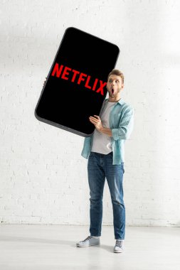 KYIV, UKRAINE - 21 Şubat 2020: Beyaz tuğla duvarın yanında netflix uygulamalı akıllı telefonun modelini tutan heyecanlı adam