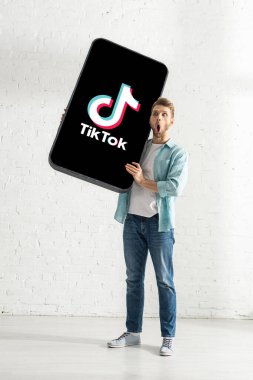 KYIV, UKRAINE - FEBRUARY 21, 2020: Surprised man with TikTok app on big model of smartphone standing at home 