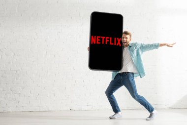 KYIV, UKRAINE - 21 Şubat 2020: Beyaz tuğla duvarın yanında netflix uygulamalı akıllı telefonun büyük modelini tutan neşeli adam