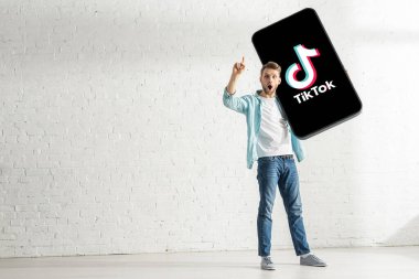 KYIV, UKRAINE - FEBRUARY 21, 2020: Surprised man showing solution gesture while holding model of smartphone with TikTok app at home  clipart