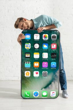 KYIV, UKRAINE - 21 Şubat 2020: Genç adam beyaz tuğla duvarın yanında iPhone ekranlı akıllı telefonun büyük modelini öpüyor
