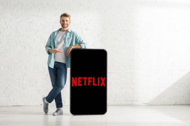 KYIV, UKRAINE - 21 Şubat 2020: Beyaz tuğla duvarın yanında netflix uygulaması olan akıllı telefonun büyük modelini gösteren neşeli adam