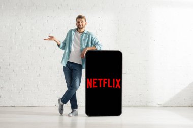 KYIV, UKRAINE - 21 Şubat 2020: Netflix uygulamalı akıllı telefon modelinin yanında omuz silkme hareketi gösteren gülen adam 