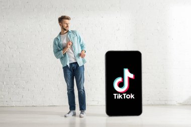 KYIV, UKRAINE - 21 Şubat 2020: Evde TikTok uygulaması ile akıllı telefonun büyük bir modelinin yanında duran kafası karışmış adam 