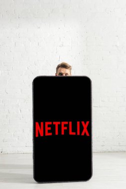 KYIV, UKRAINE - FEBRUARY 21, 2020: Man looking at camera near big model of smartphone with netflix app at home  clipart