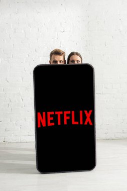 KYIV, UKRAINE - 21 Şubat 2020: Evinde netflix uygulaması olan dev akıllı telefon modelinin yakınındaki kameraya bakan çift 