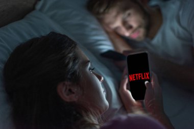 KYIV, UKRAINE - 21 Şubat 2020: Netflix uygulamalı akıllı telefonu elinde tutan kızın seçici odak noktası gece yatakta erkek arkadaşının yanında 