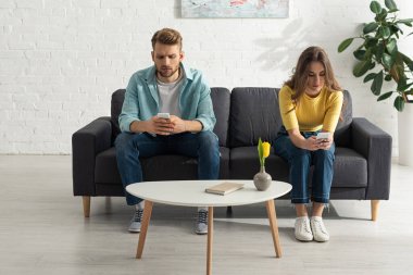 Young couple using smartphones on couch near book on coffee table  clipart