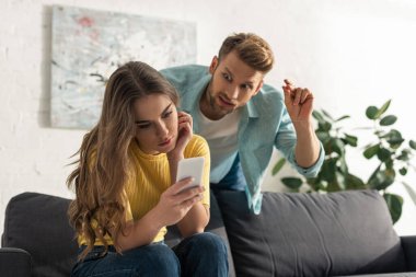 Akıllı telefonun seçici odağı oturma odasındaki kızgın erkek arkadaşın yanındaki koltukta oturan kıza bağlıydı.