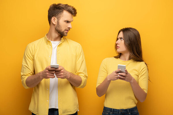 Confused couple with smartphones looking at each other on yellow background
