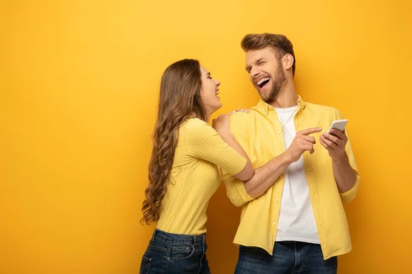 Laughing Girl Embracing Boyfriend Pointing Smartphone Yellow Background — Stock Photo, Image