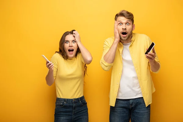 Surpreendido Jovem Casal Segurando Smartphones Olhando Para Câmera Fundo Amarelo — Fotografia de Stock