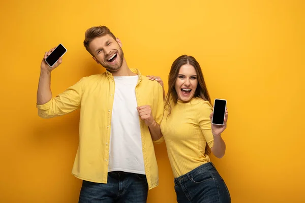Vrolijk Jong Stel Tonen Smartphones Kijken Naar Camera Gele Achtergrond — Stockfoto
