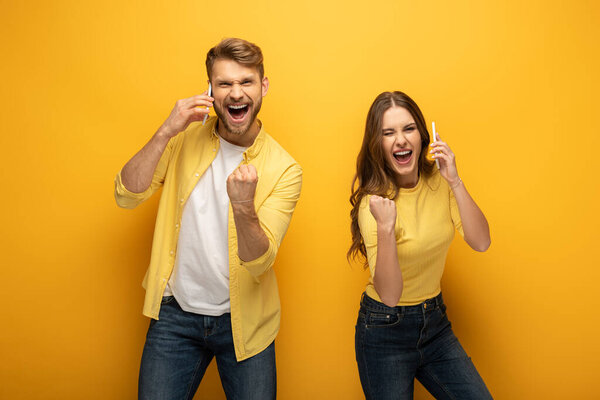 Cheerful couple showing yes gesture while talking on smartphones on yellow background