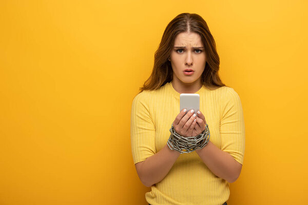 Confused woman with metal chain around hands holding smartphone and looking at camera on yellow background