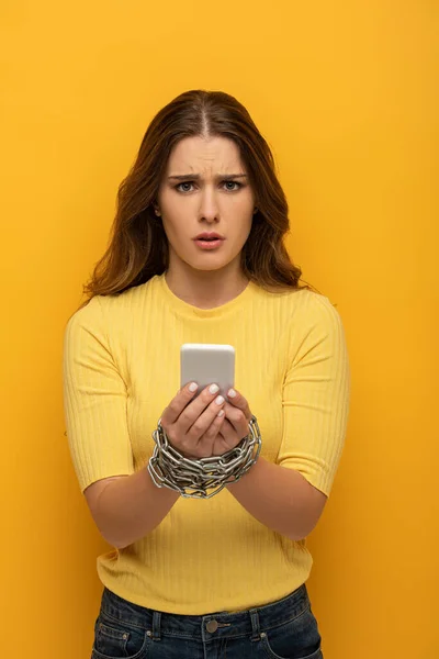 困惑的女孩一边拿着智能手机一边用黄色背景的金属链捆着手看着相机 — 图库照片