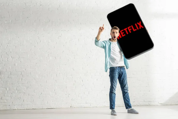 stock image KYIV, UKRAINE - FEBRUARY 21, 2020: Shocked man having idea while holding big model of smartphone with netflix app at home 