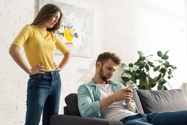 怒气冲冲的女人手放在屁股上 站在智能手机旁边 男人靠在沙发上 — 图库照片