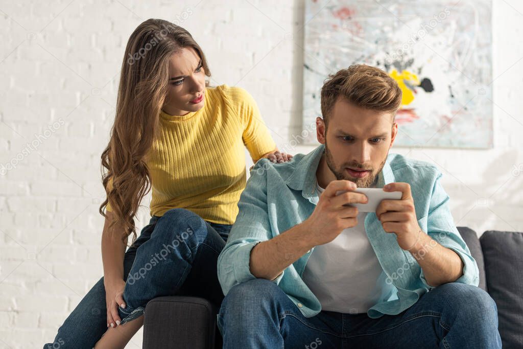 Worried girl sitting on couch near smartphone depended boyfriend 