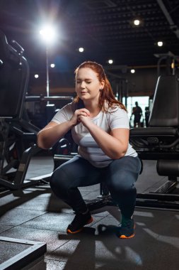 concentrated overweight girl with closed eyes squatting with clenched hands in gym clipart