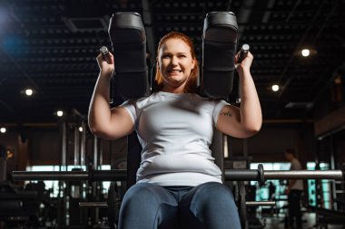 cropped view of purposeful overweight girl doing arms extension exercise on fitness machine clipart