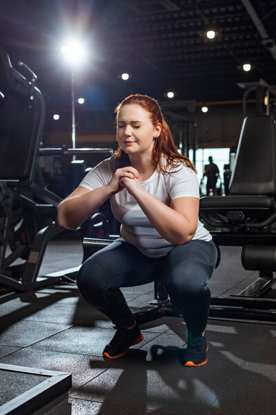 concentrated overweight girl with closed eyes squatting with clenched hands in gym