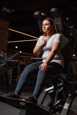 confident overweight girl looking away while sitting on fitness machine clipart