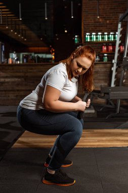 Spor salonunda ağırlık diskiyle çömelmiş kilolu bir kız.