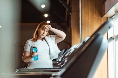 selective focus of tired overweight girl touching forehead and holding sports bottle while standing at treadmill clipart