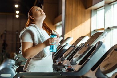 tired overweight girl with towel on shoulder holding sports bottle and looking up while standing on treadmill clipart