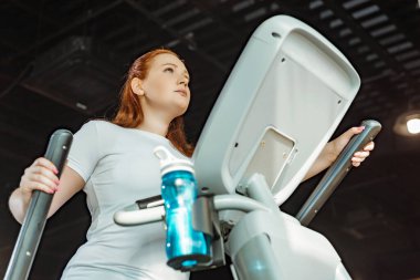 Düşük açılı, odaklanmış, kilolu bir kızın spor salonunda stepper üzerinde çalışması.
