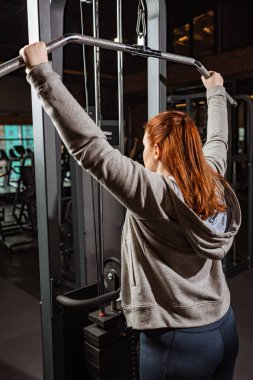 Gri kapüşonlu şişman kız spor makinesinde kol egzersizi yapıyor.