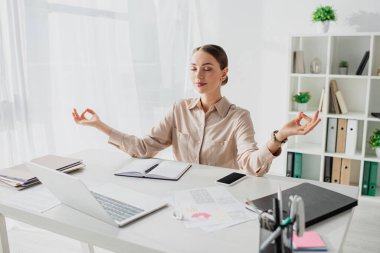 Mutlu iş kadını gözleri kapalı meditasyon yapıyor ve iş yerinde bilgisayarı ve akıllı telefonu olan Gyan Mudra