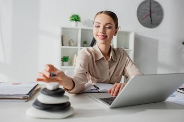 Zen taşlarıyla iş yerinde dizüstü bilgisayarla çalışan gülümseyen iş kadınının seçici odağı