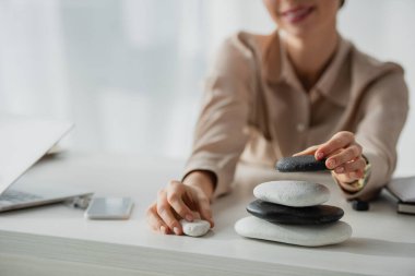 cropped view of businesswoman sitting at workplace with zen stones, smartphone and laptop  clipart