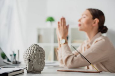 İş kadını seçici bir şekilde meditasyon yapıyor. İş yerinde Buda kafası ve tütsü çubuğuyla namaste jesti yapıyor.