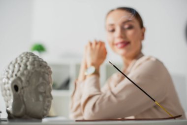 selective focus of happy businesswoman sitting in office with Buddha head and incense stick clipart